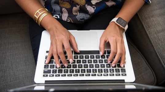 Aerial shot of dark-skinned person with pink fingernail polish wearing an Apple watch typing on an Apple laptop