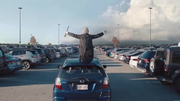 Geico TV ad with a person riding on top of car in a parking lot